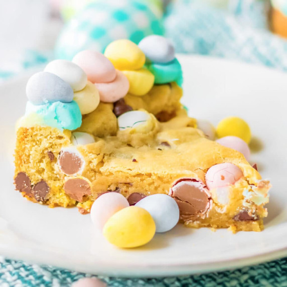 Slice of chocolate mini egg cookie cake on a plate decorated with more candy Easter eggs.