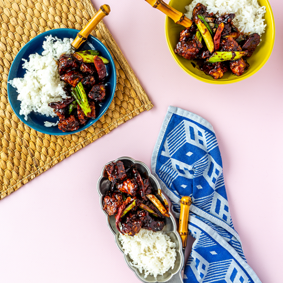 Three plates of Mongolian tempeh clusters with rice and bamboo cutlery on a soft pink backdrop.