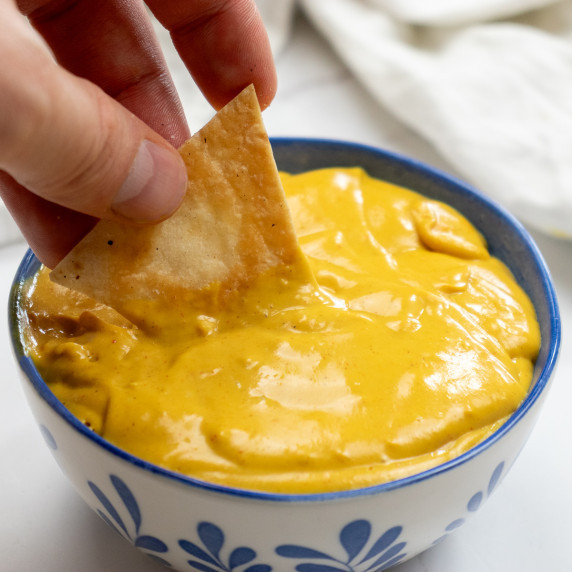 My hand dipping a homemade nacho into the gooey vegan nacho cheese