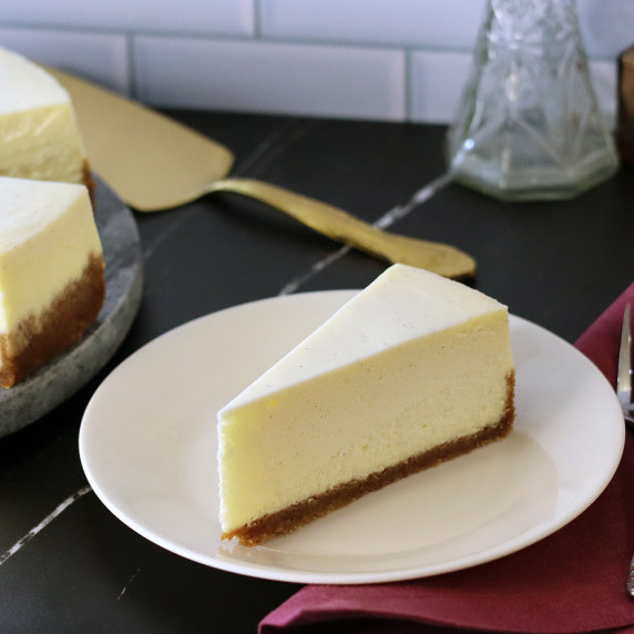A slice of New York style cheesecake on a white plate on a black countertop.