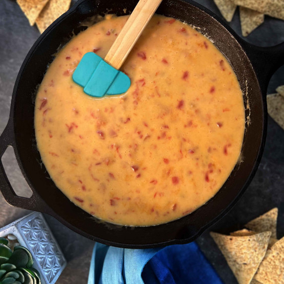 Nacho cheese dip in cast-iron skillet with teal rubber spatula in the cheese on counter top.