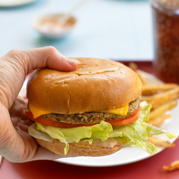 A burger being held in front of a plate of fries and a glass of soda.