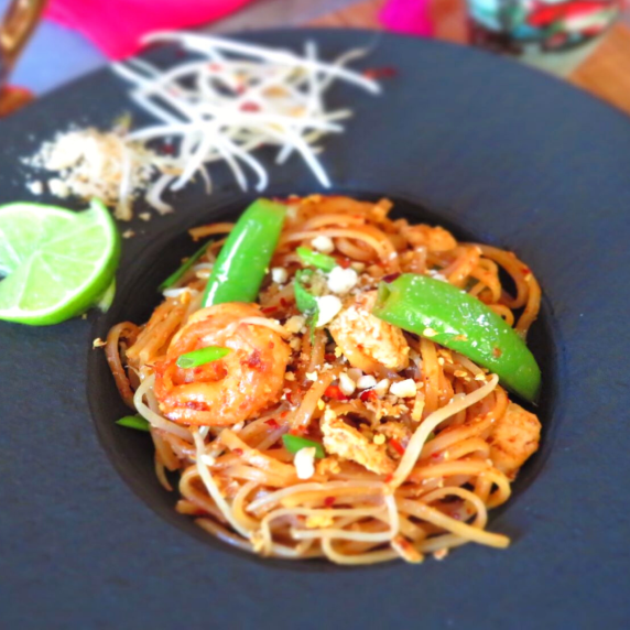 Pad thai with shrimp, snap peas, chili flakes, and crushed peanuts in a black bowl