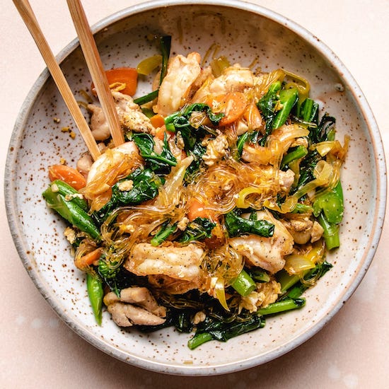 Stir fried chicken, shrimp, glass noodles, carrots and broccolini in stoneware bowl with chopsticks