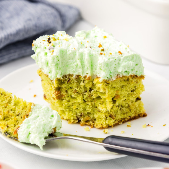 A slice of green frosted pistachio pudding cake on a plate with a bite removed.