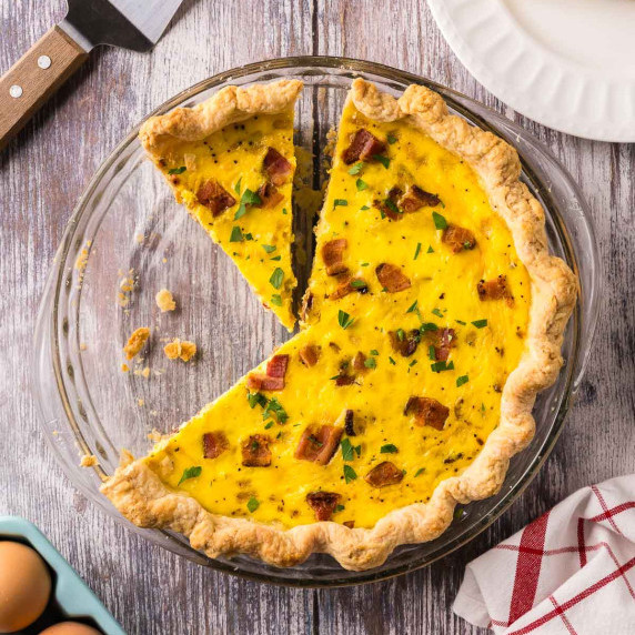 A quiche with three slices cut, two of which have been removed.  topped with bacon