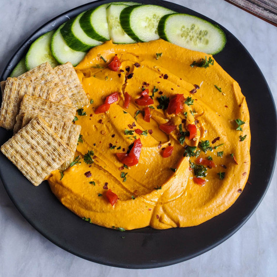 Bright orange hummus with green cukes on a black plate against a marble background.