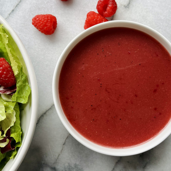 Raspberry vinaigrette dressing in a white bowl on a marble countertop with fresh raspberries.