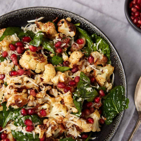 A bowl filled with roasted cauliflower, fresh spinach leaves, grated cheese, and pomegranate seeds