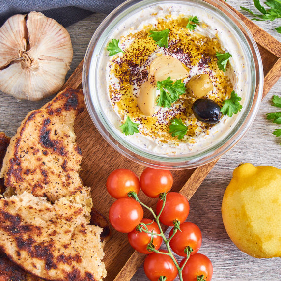 A bowl of Roasted Garlic Lemon Labneh surrounded by flatbread, tomatoes, garlic and lemon