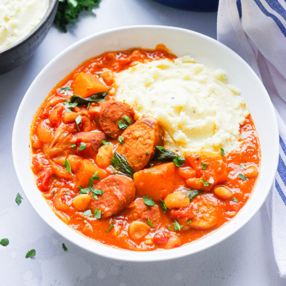 Sausage and bean casserole in a white bowl, served with mashed potatoes and garnished with parsley.