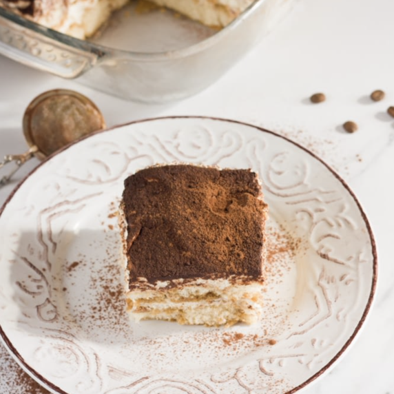 a slice of tiramisu on a white and brown plate on a white countertop with a pan of tiramisu 