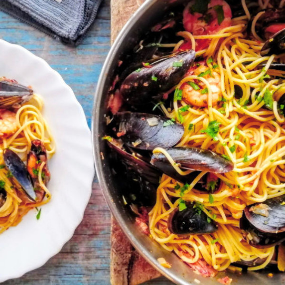 A large steel pot sits filled with shrimp and mussels pasta waiting to be served to a plate nearby