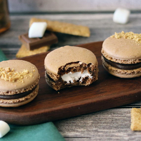 Three s'mores macarons on a wooden serving tray with a bite taken from the center macaron.