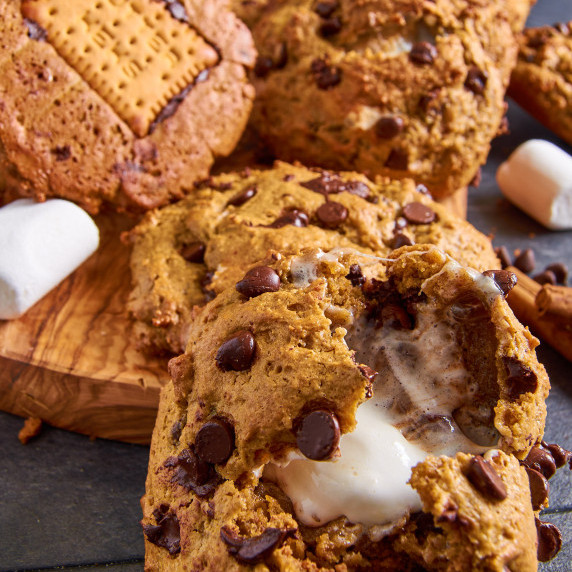 A wooden board full of Sourdough Pumpkin S’mores Cookies, one broken open, showing gooey marshmallow