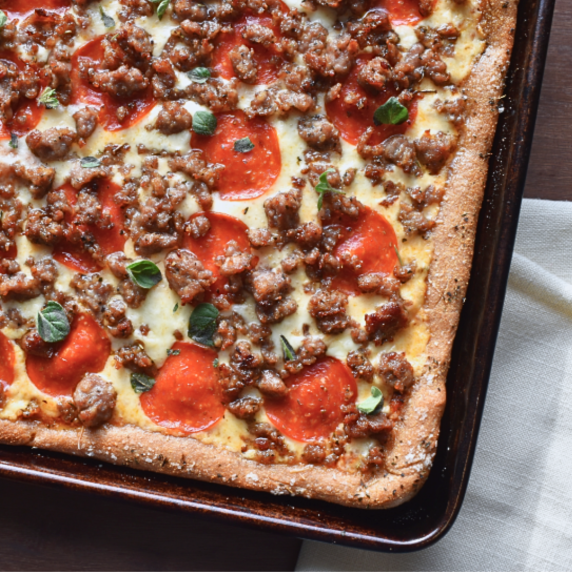 A pizza topped with sausage, pepperoni, cheese, and fresh oregano in a sheet pan on a dark table