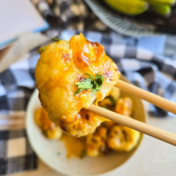 Wooden chopsticks holding a cauliflower floret dredged in an orange sauce.