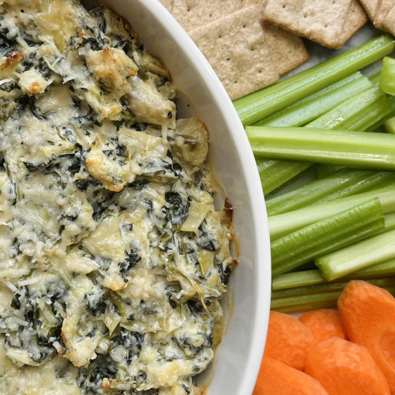 White baking dish filled with spinach artichoke dip alongside carrots, celery and crackers.