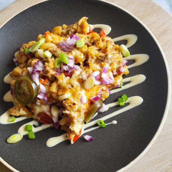 A heap of cheesy mac on a grey plate with a white sauce drizzle and colourful garnishes