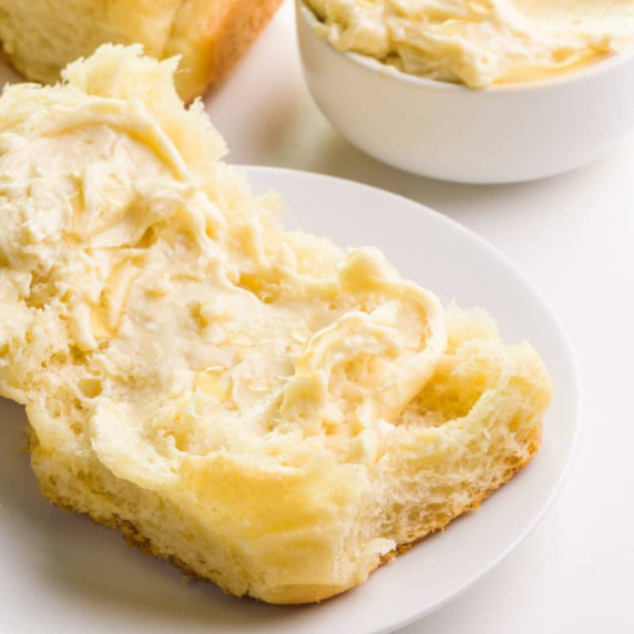 A bun sits on a plate and is covered with vegan honey butter. There's more butter in a bowl.