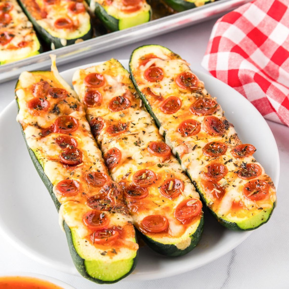 3 zucchini pizza boats on a plate topped with mini pepperoni with more on a pan in the background.