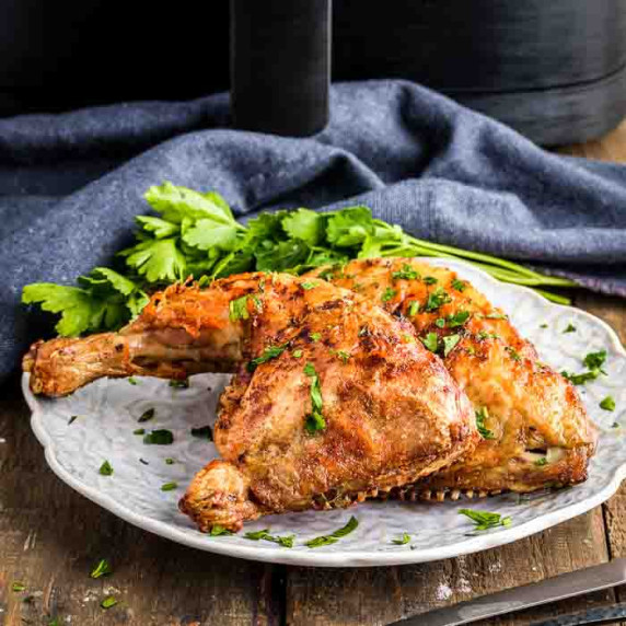 Side shot of two chicken quarters garnished with herbs on a white plate on a wooden table.