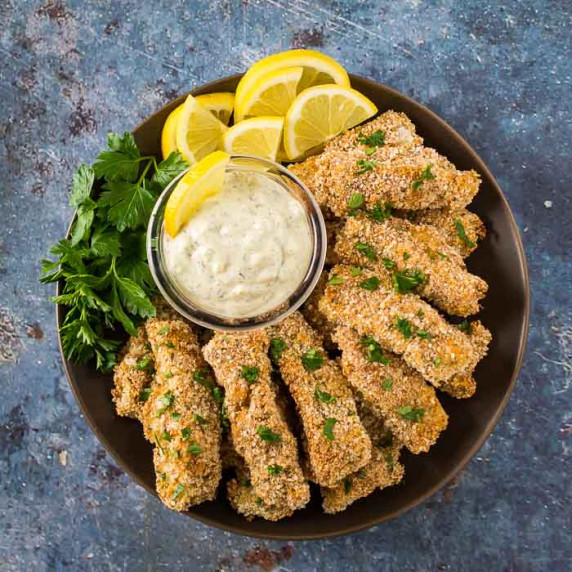 Side angled shot of breaded fish sticks with tartar sauce, lemon wedges, and herbs on the side.