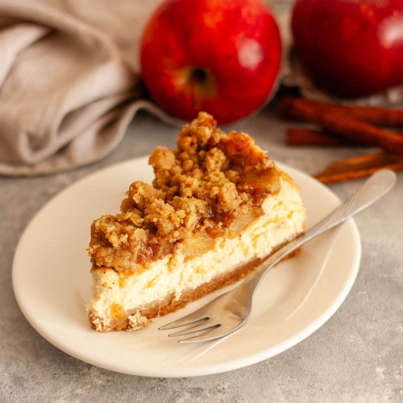A slice of apple crumble cheesecake on a white plate next to apples and cinnamon sticks.
