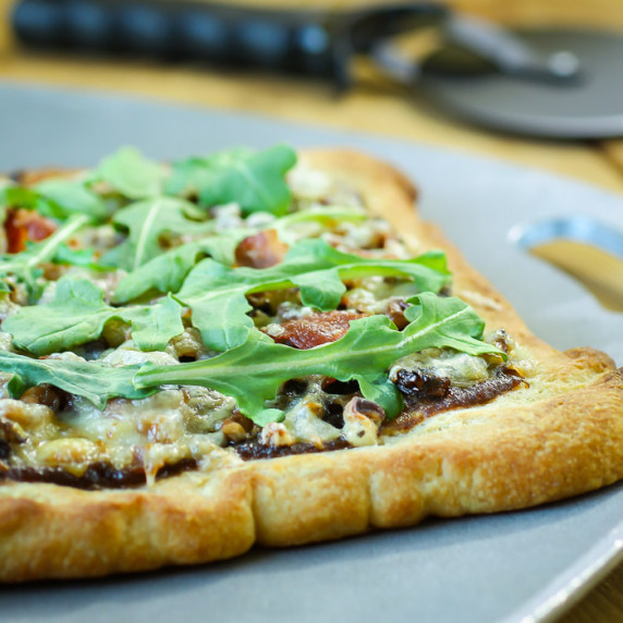 Side angled view of the whole onion bacon pizza on a grey cutting board on a wooden surface.