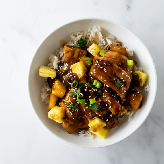 teriyaki chicken with pineapple over rice in a white bowl topped with sesame seeds and green onion.