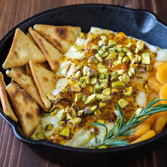 Side view of baked brie with jam in a skillet with herbs and pita chips.