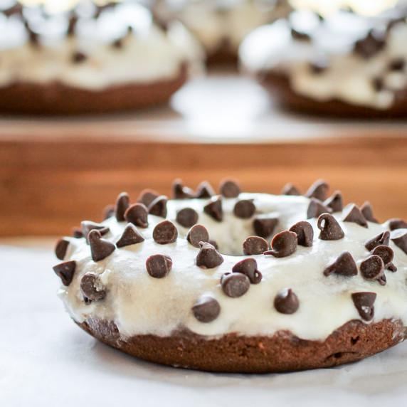 Side shot of a chocolate donut with chocolate chips on a white surface with more donuts in the backg