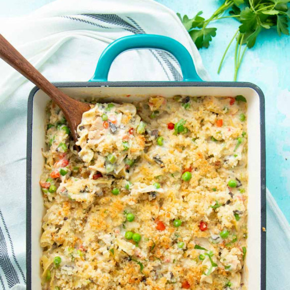 Overhead of a wooden spoon scooping tuna noodle casserole from a baking dish.