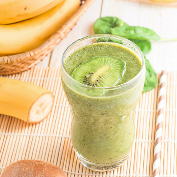 Side shot of kiwi smoothie with a slice of kiwi on top in a clear glass on a bamboo mat.