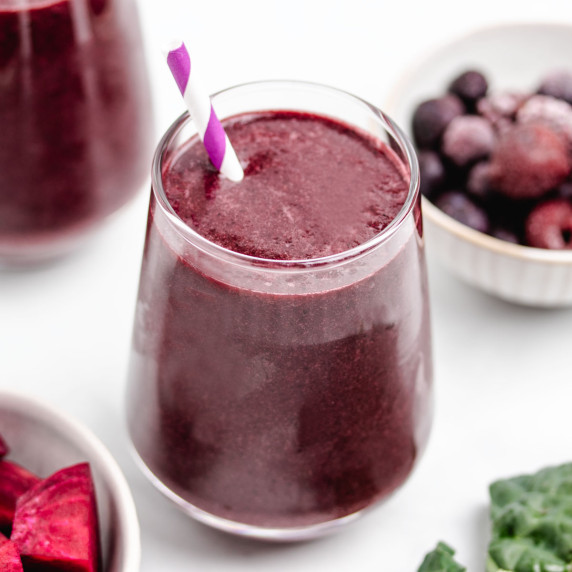 A kale beet smoothie in a glass with a straw and frozen berries in the background.