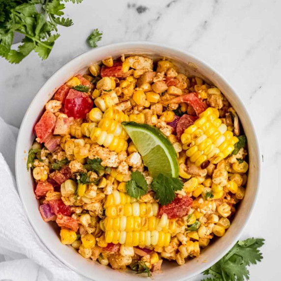 Top view of a white bowl filled with colorful corn salad garnished with a lime wedge and cilantro.