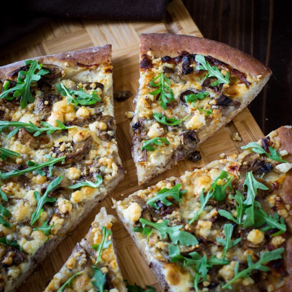 Side angled shot of multiple slices of steak pizza garnished with arugula on a wooden cutting board.