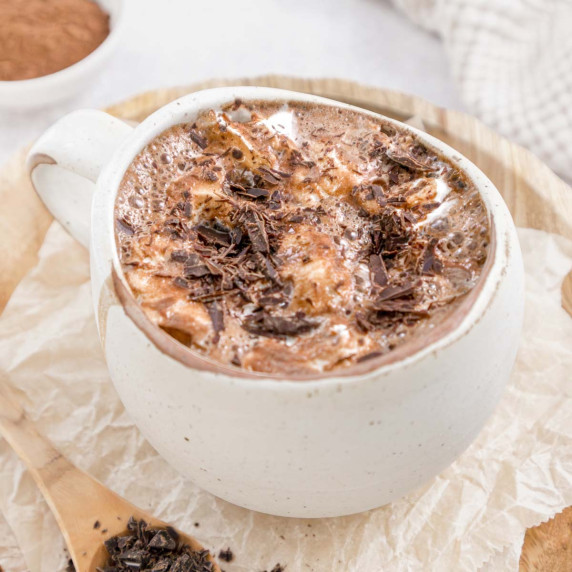 Hot chocolate topped with flaked chocolate in a cream mug sitting on wooden plate.