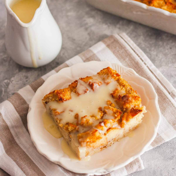 Bread pudding with vanilla sauce on a brown and white towel next to a jug of vanilla sauce.