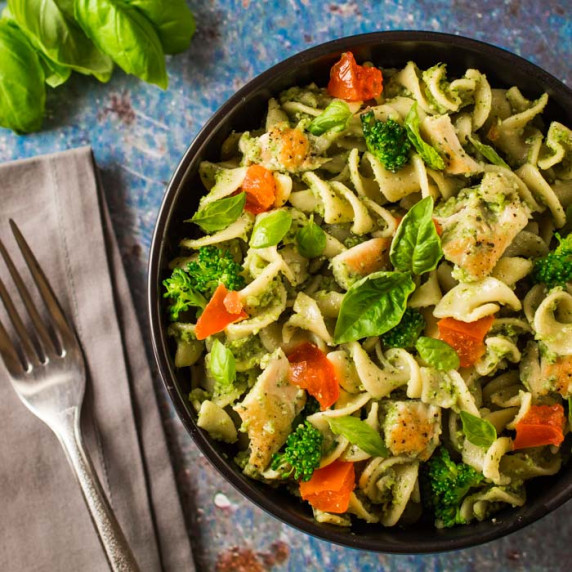 Overhead shot of chicken pesto pasta garnished with basil in a black bowl on a blue surface.