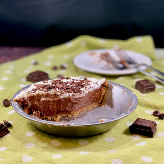 Half chocolate cream pie in the pan with 2 forks on a plate in background on a green polka dot towel