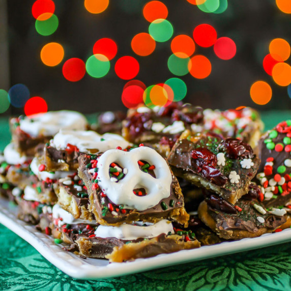 Side view of Christmas Bark on a white square plate on a green cloth with Christmas lights behind. 