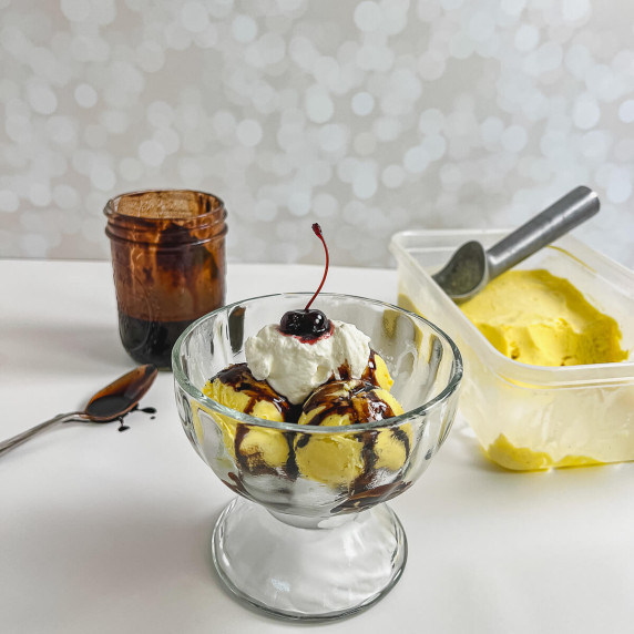French vanilla ice cream sundae in front of jar of chocolate syrup, spoon, and tub of ice cream.