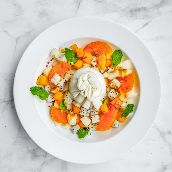 Overhead shot of colorful burrata salad garnished with mint in a white bowl on a marble table.