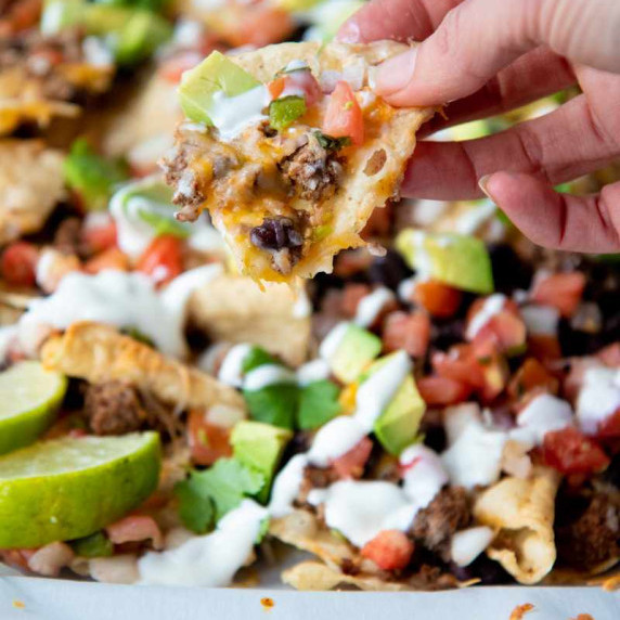 A hand holds up a loaded nacho chip above a platter of loaded ground beef nachos.