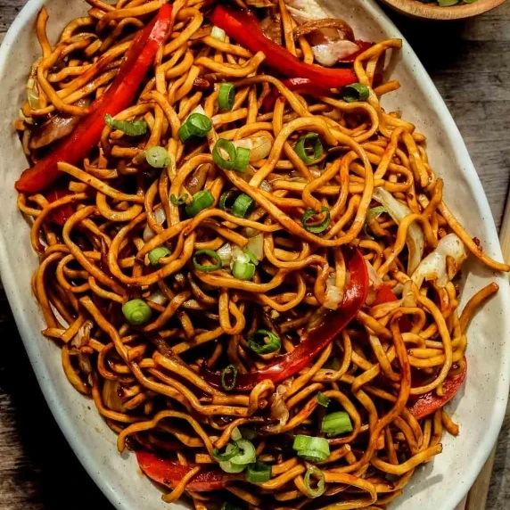 A plate of Hakka noodles on a white plate with green onions on top.