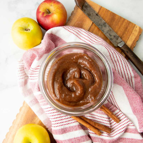 Top view of apple butter swirled in a glass bowl with fresh apples and whole cinnamon sticks around.
