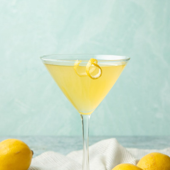 A limoncello martini in a glass with a lemon twist on the rim and whole lemons around the glass.