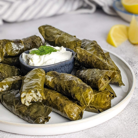 Stuffed grape leaves on a plate next to a bowl with yogurt and lemon slices.