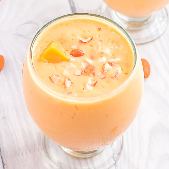 Overhead shot of mango lassi in a clear curvy glass garnished with mango chunks and pieces of almond